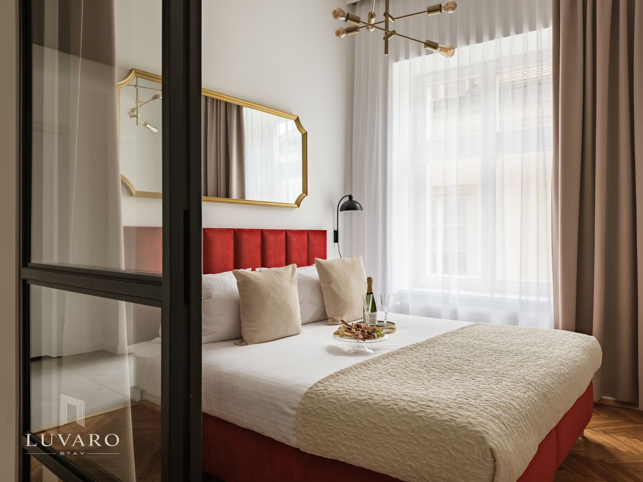A hotel room with a bed, a bedside table, a mirror, a chandelier, and a window with curtains.