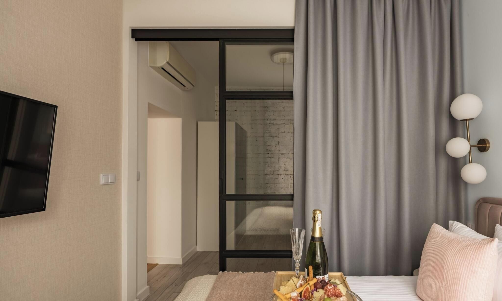 A hotel room with a bed, a TV mounted on the wall, a window with curtains, and a bedside table with a bottle and glasses.