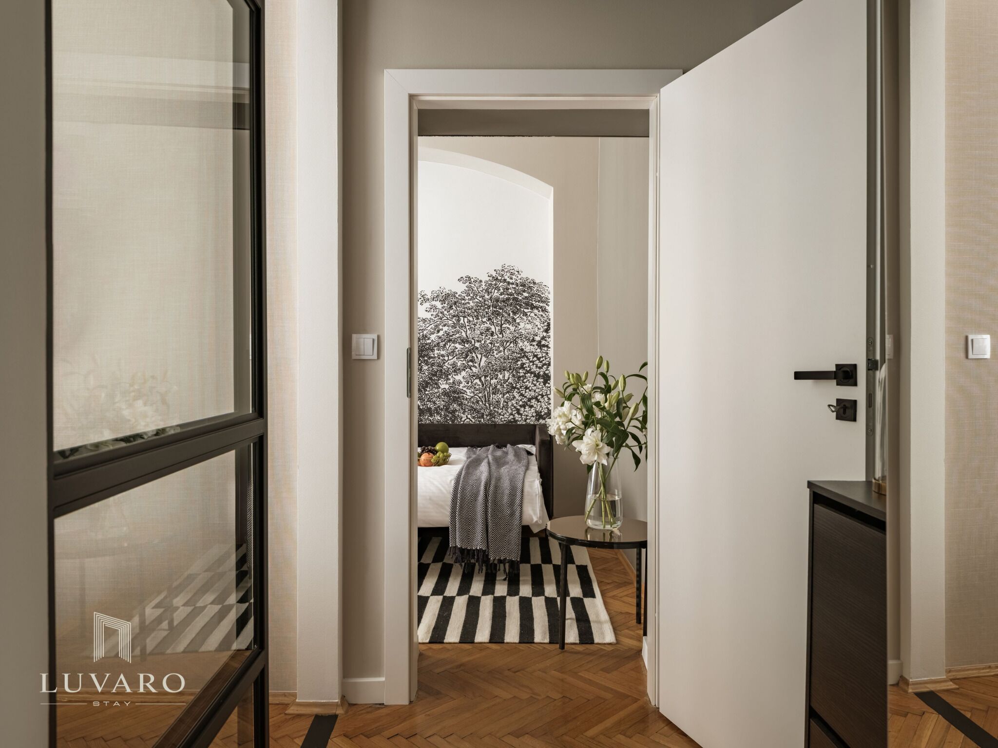 Ein Hotelzimmer mit Schachbrettboden, einem Bett mit schwarz-weiß gestreifter Decke, einer Vase mit Blumen und einer Wand mit einem schwarz-weißen Baumdekor.