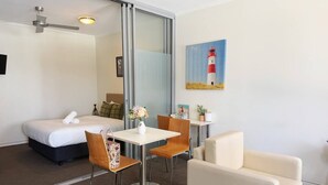 Hotellihuone, jossa on sänky, ruokapöytä tuoleineen, sohva, majakan kuva ja kattotuuletin.