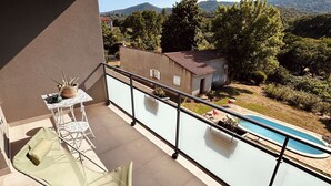 Un balcón con vista a la piscina, muebles de exterior y un edificio pequeño al fondo.
