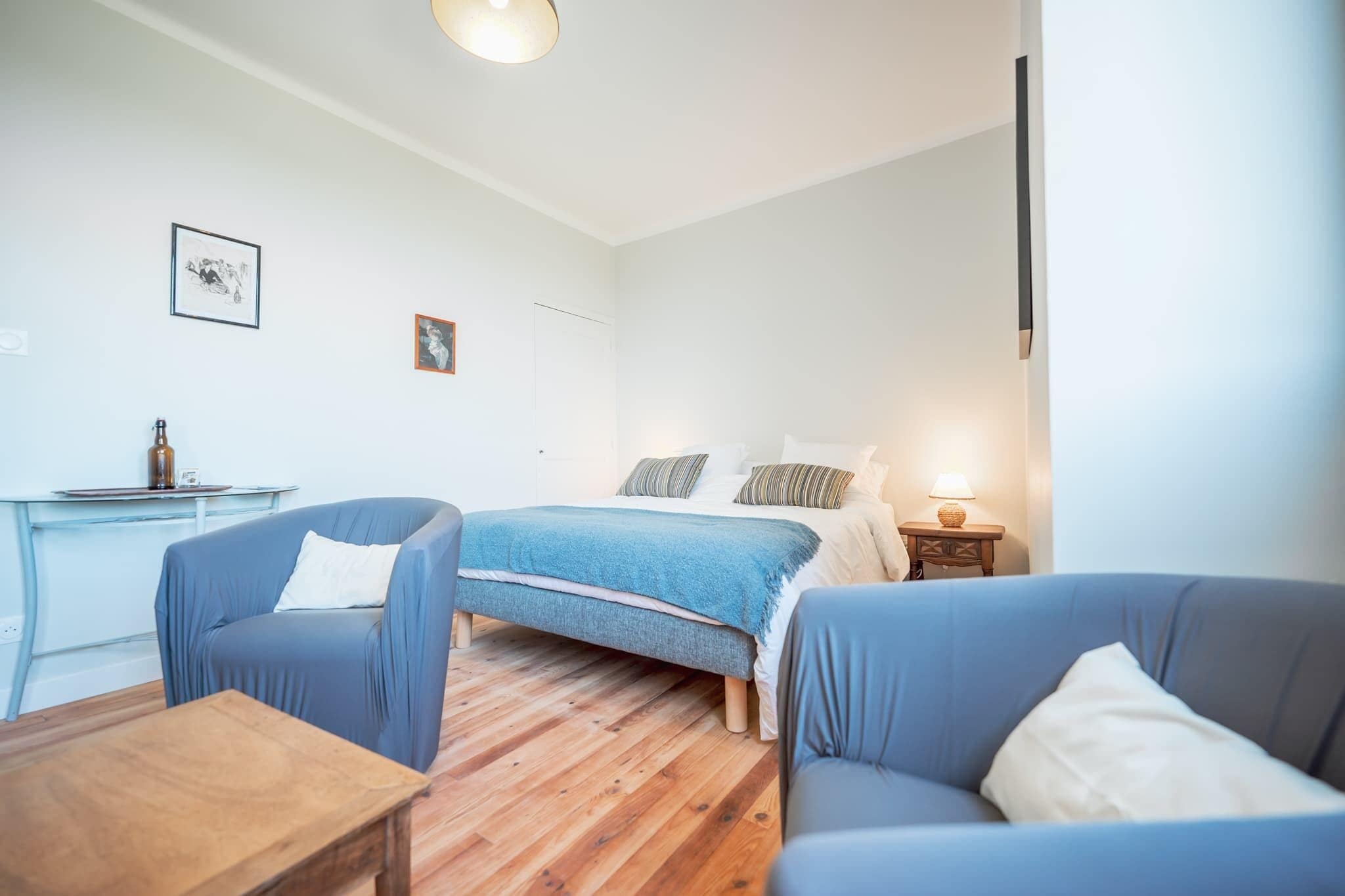Une chambre à coucher avec un lit, deux fauteuils bleus, une table en bois, un bureau avec une lampe et un petit tableau accroché au mur.