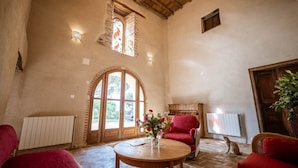 A rustic interior with a piano, red armchairs, a wooden table, and a large arched window. - Les Jardins de la Longagne (Saussenac)