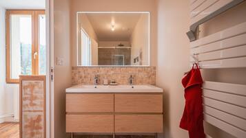 A bathroom with a double sink, a mirror with a light, a towel rack, and a shower area.