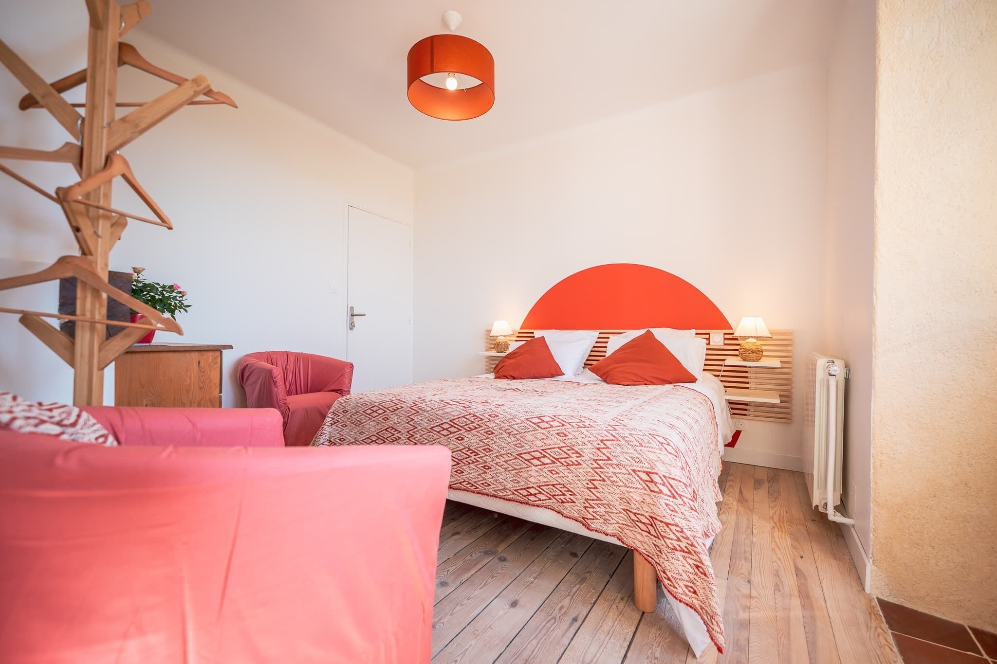A bedroom with a bed, a wooden chair, a hanging chair, a bedside table with a lamp, and a radiator.