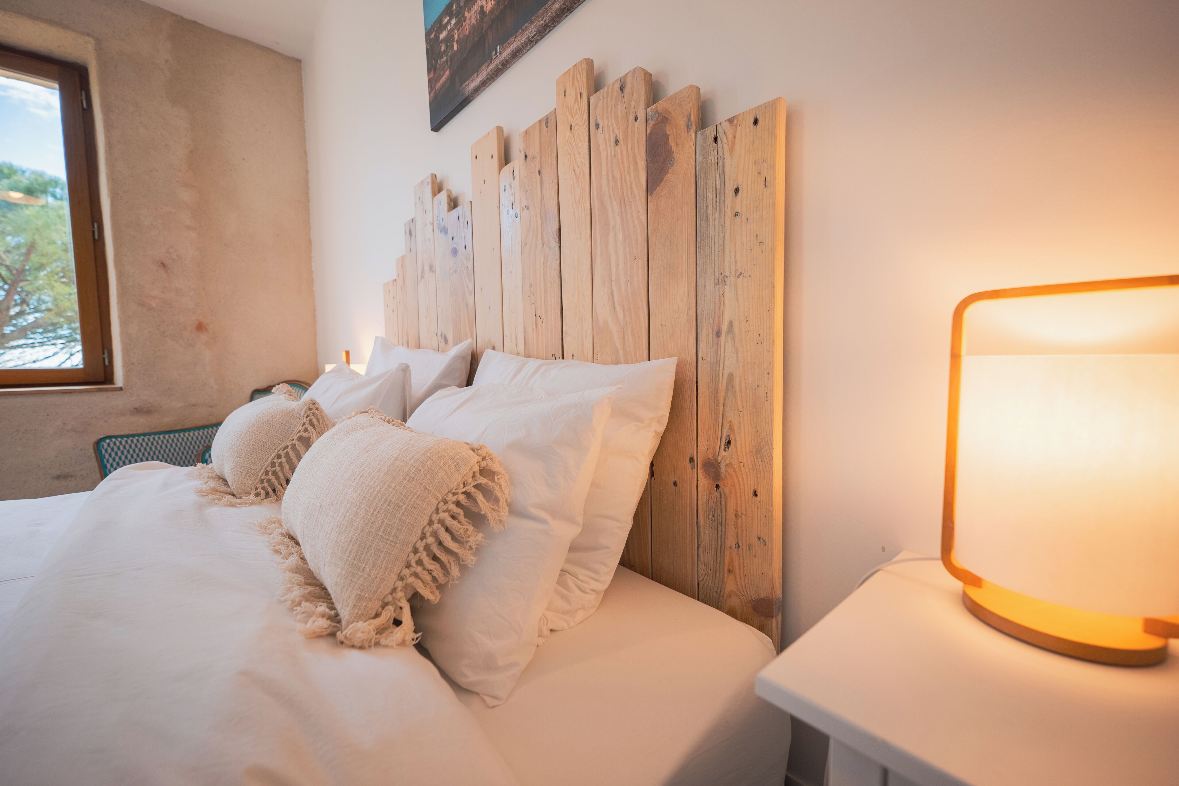 Een bed met witte lakens en een houten hoofdbord, een nachtkastje met een lamp, en een raam met uitzicht op bomen.