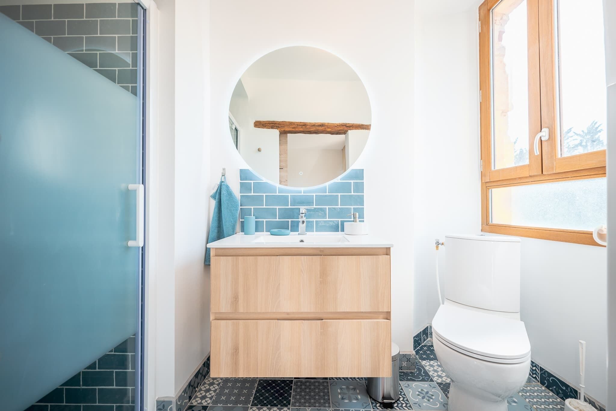 Une salle de bain avec une porte bleue, un miroir rond, des toilettes blanches et un lavabo surmonté d’une serviette bleue.