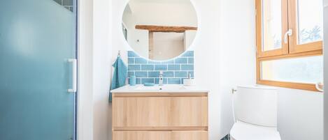 Une salle de bain avec une porte bleue, un miroir rond, des toilettes blanches et un lavabo surmonté d’une serviette bleue.