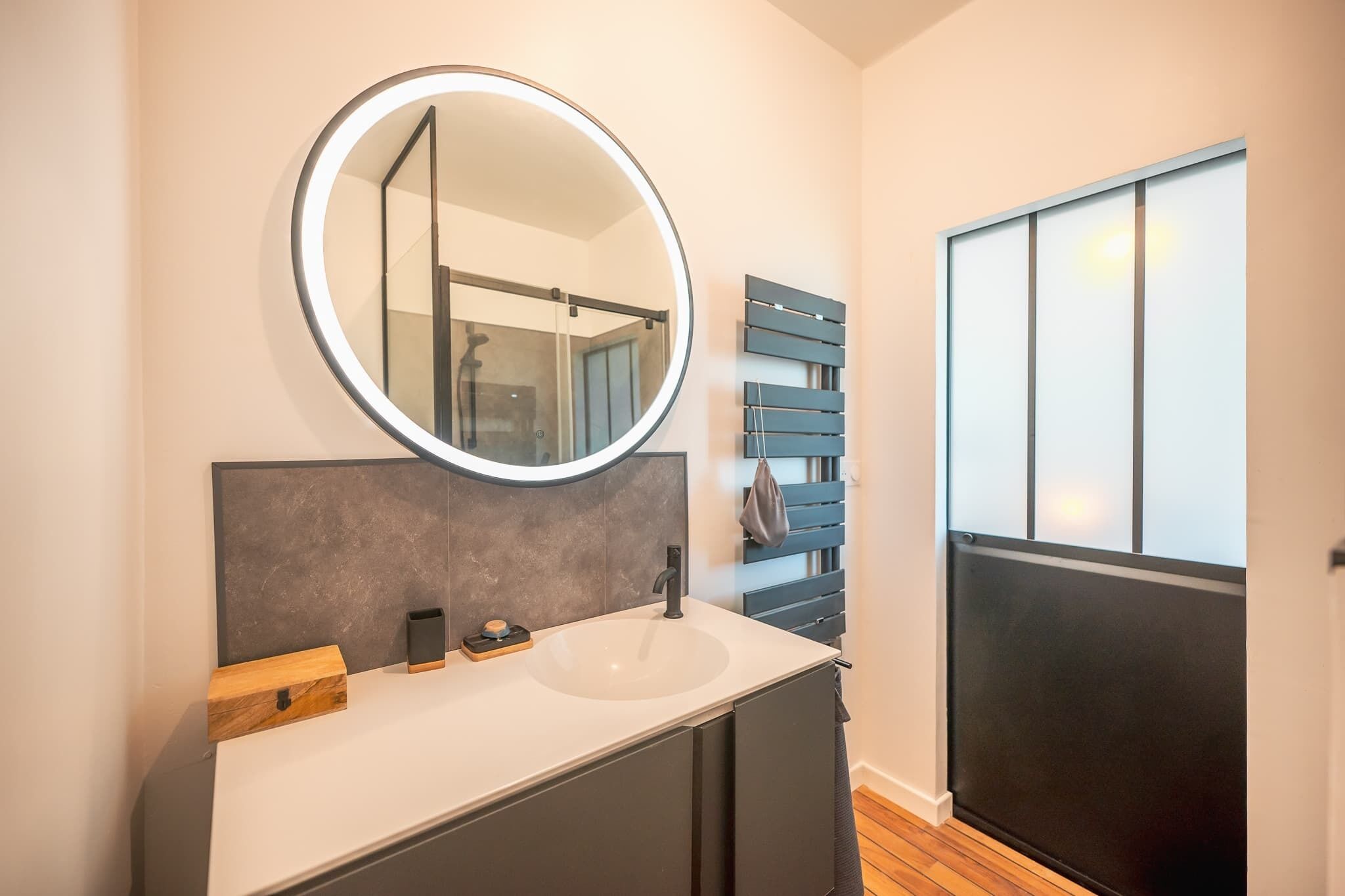 A modern bathroom with a round mirror, a white sink, dark cabinets, and a wooden bench.