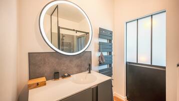 A modern bathroom with a round mirror, a white sink, dark cabinets, and a wooden bench.