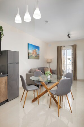 A modern living room with a glass dining table, grey chairs, a grey sofa, a painting on the wall, and a kitchen area with a fridge. - Edka Suites - Modern Sunlit 2BD Apartments with Balconies - Close to Sliema Promenade, St Julian's & St Georges Bay (Gzira)