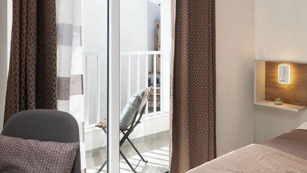 Moderni hotellihuone, jossa on sänky, tuoli, parveke, jossa on myös tuoli, sekä seinään kiinnitetty valaisin.