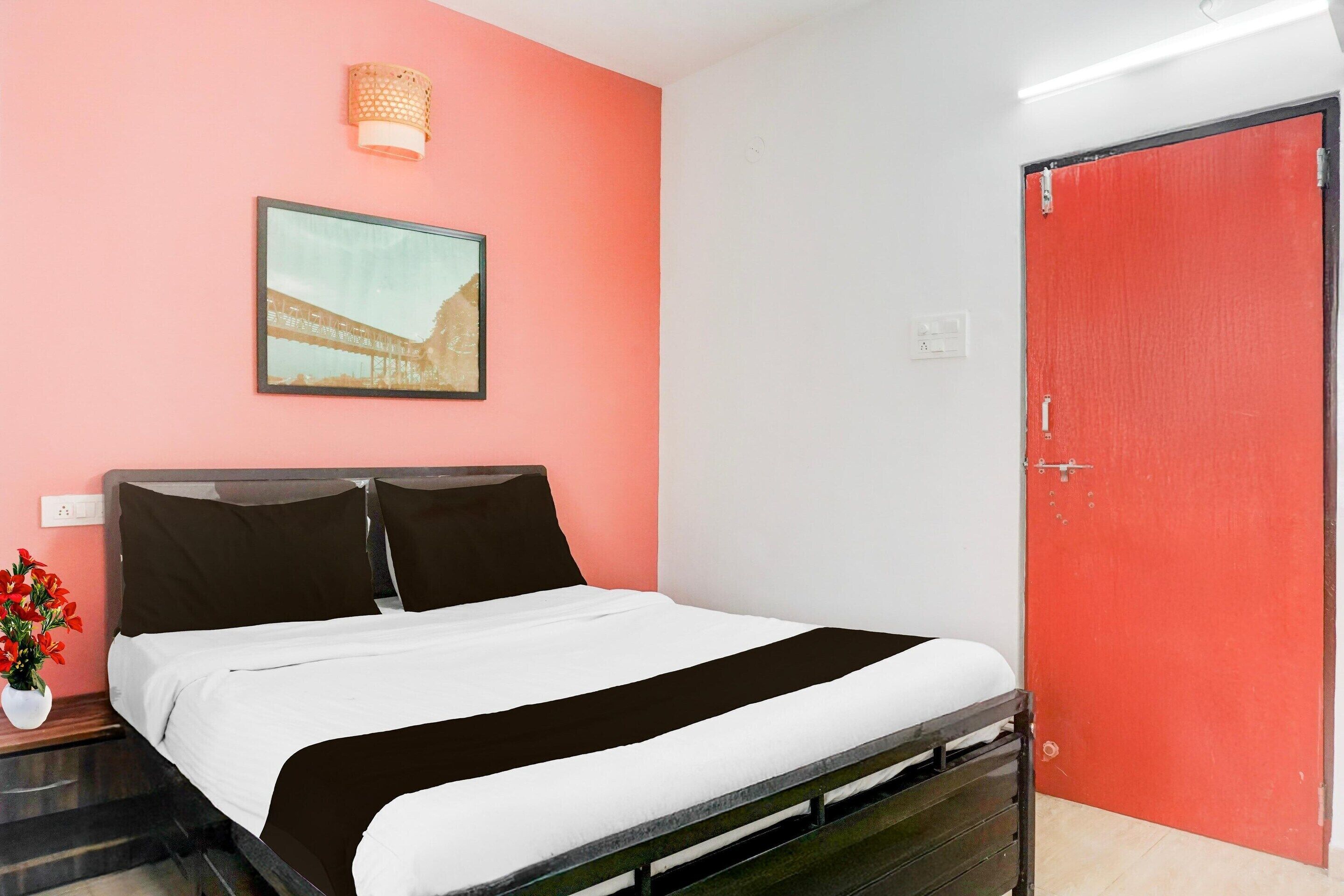 A bedroom with a bed, a nightstand with a vase of flowers, a red door, and a framed picture on the wall.