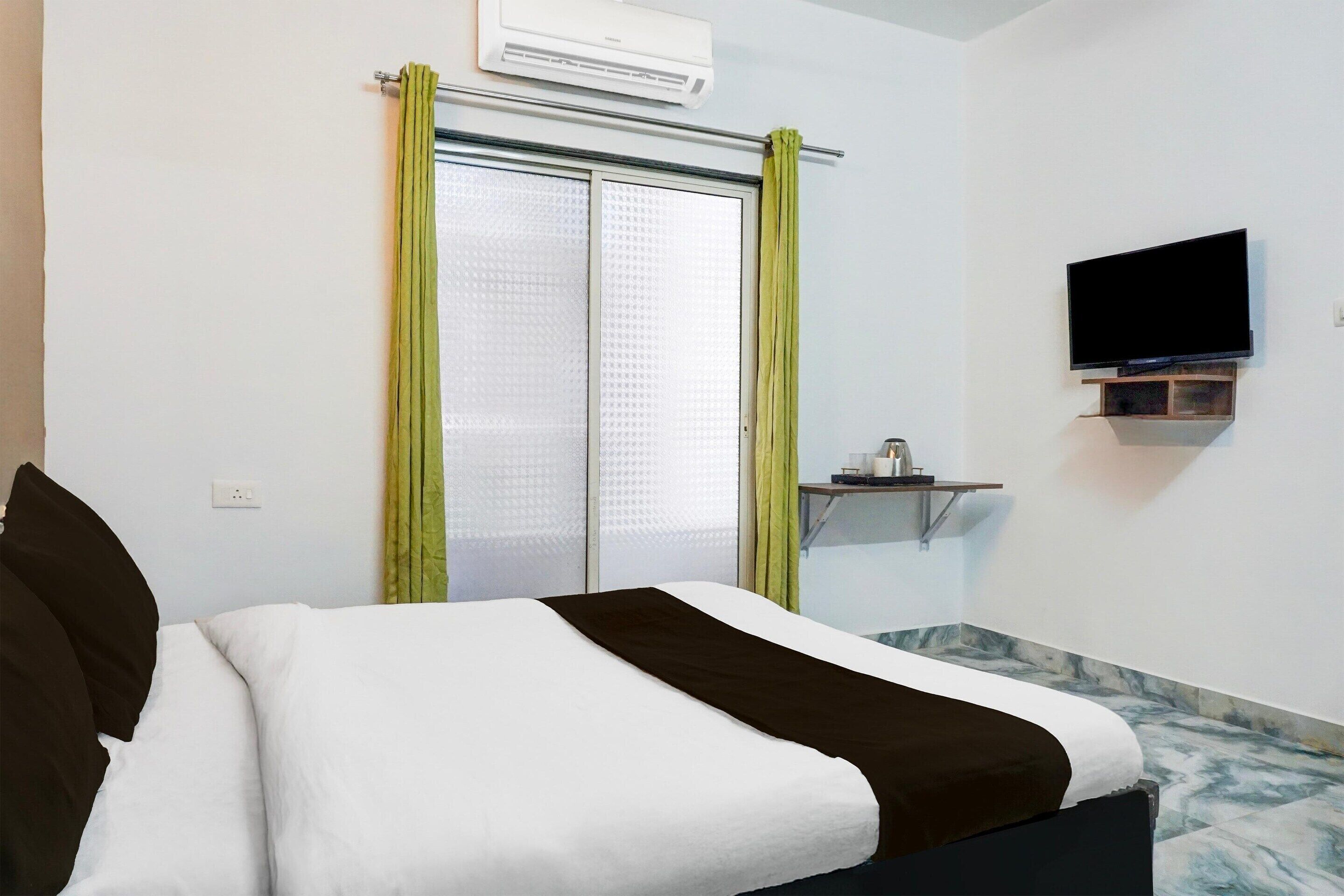 A hotel room with a bed, a television mounted on the wall, a small table with a lamp, and a window with curtains.
