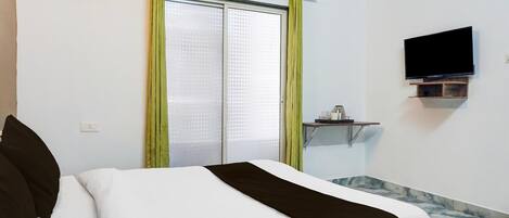 A hotel room with a bed, a television mounted on the wall, a small table with a lamp, and a window with curtains.