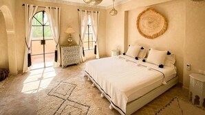 A spacious bedroom with a large bed, a round woven wall hanging, and a view through a window with curtains. - Riad Selouane (Marrakech)