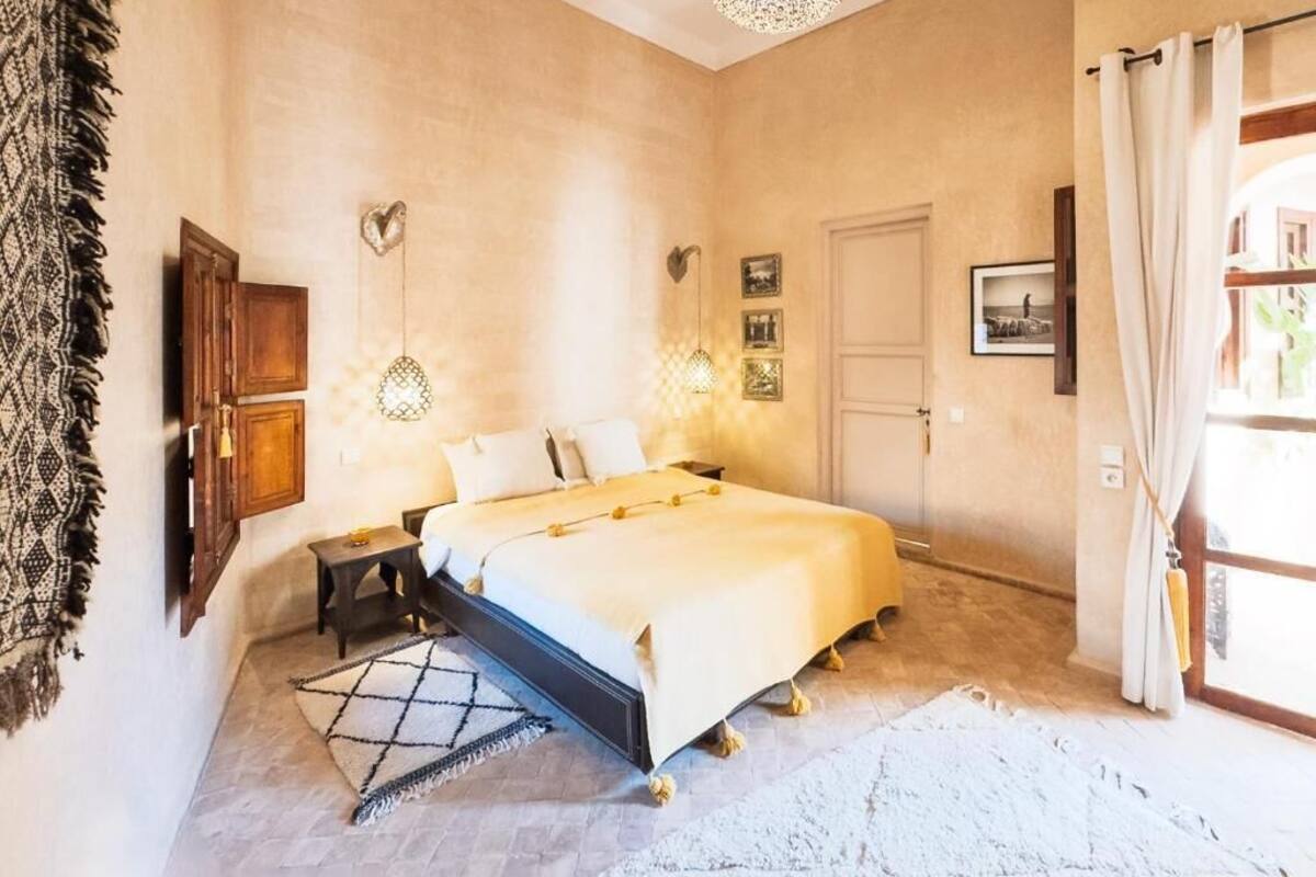 Un dormitorio con cama, mesitas de noche, lámpara de pared, una puerta y una ventana con cortinas.