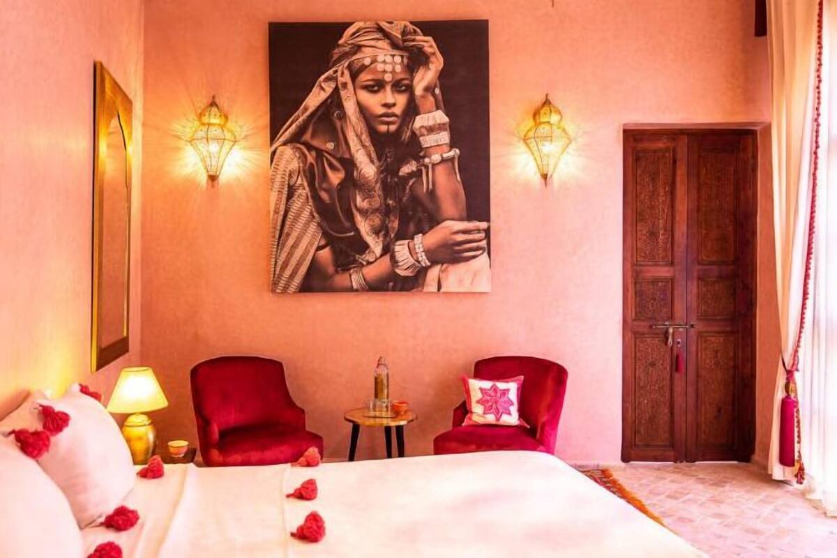 Habitación de hotel con una cama, dos sillones rojos, una mesita con una botella y un cuadro colgado en la pared.