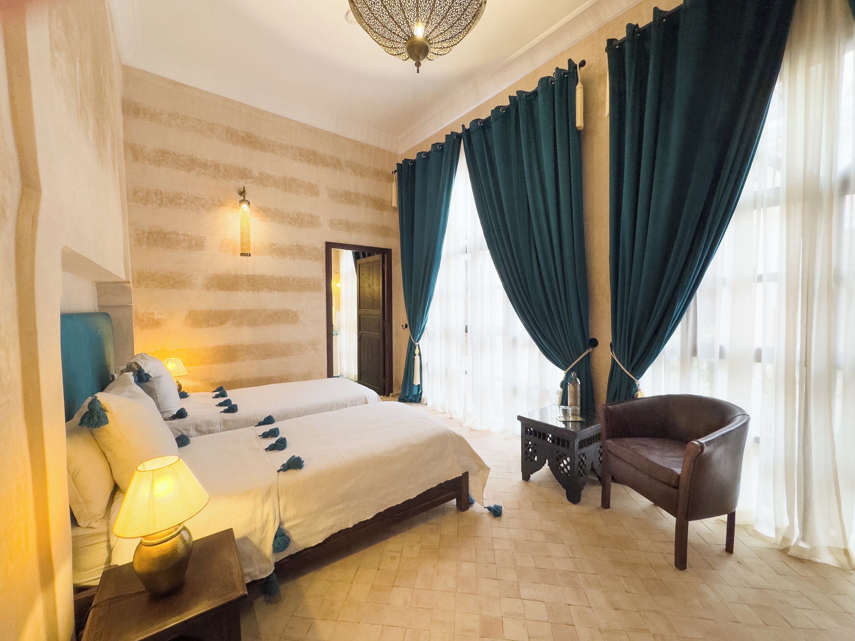 Una habitación de hotel con una cama, una silla, una mesita de noche, una lámpara y un ventanal con cortinas.