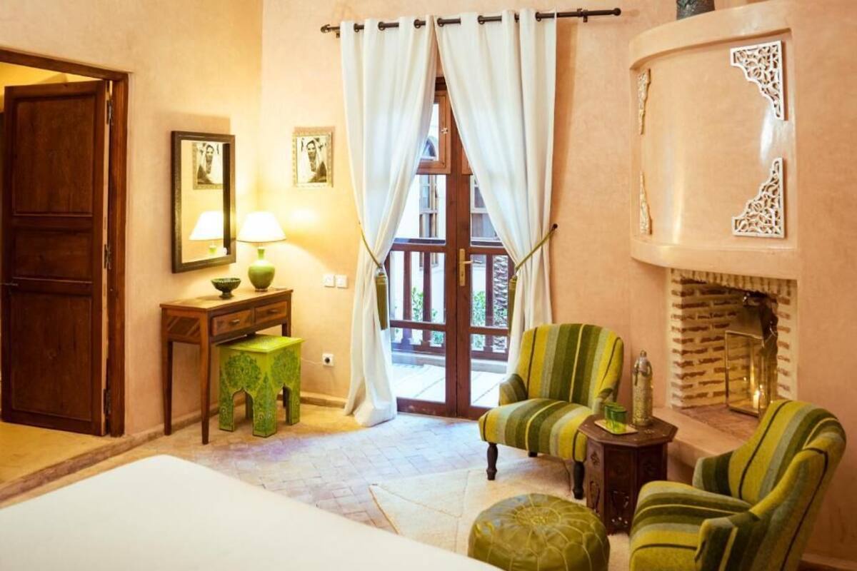 Un dormitorio con cama, una silla, una mesita con lámpara, chimenea y puerta que da a un balcón.