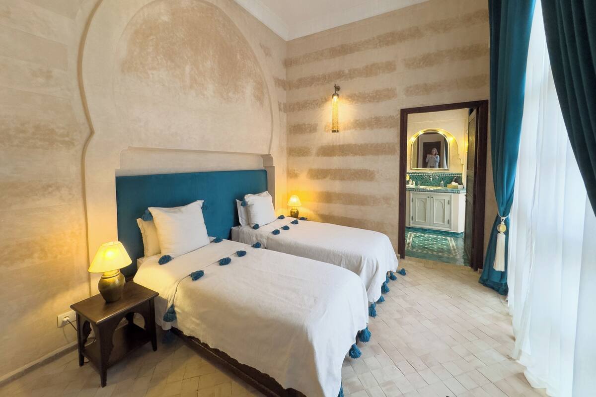 Habitación de hotel con dos camas, una mesita de noche con lámpara, un espejo y vistas a un baño con suelo de baldosas.