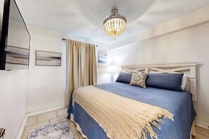 A bedroom with a bed, a chandelier, a window with curtains, and a painting on the wall. - Sunchase 203 (Gulf Shores)