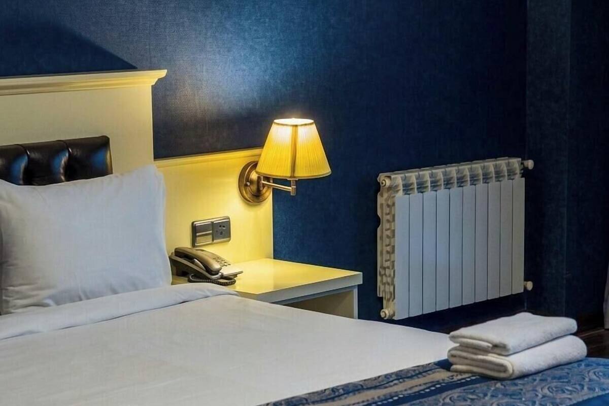 A hotel room with a bed, a bedside lamp, a telephone, a radiator, and a folded towel on the bed.