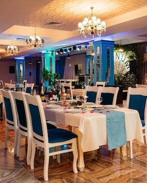 A dining area with tables set for a meal, chairs with blue cushions, and chandeliers. - Gallery & Sea Hotel (Sumqayit)