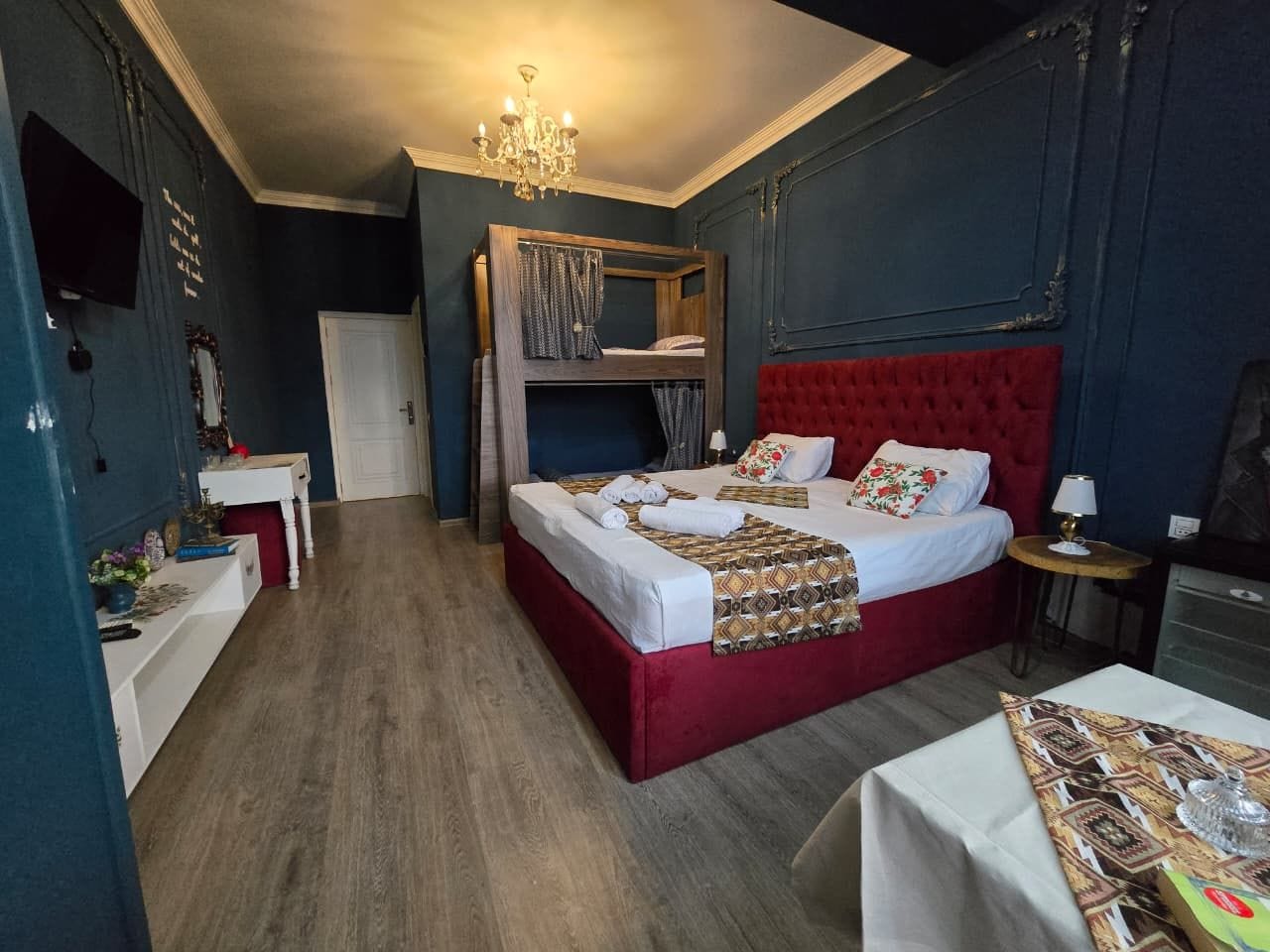 A bedroom with a red bed, a wooden bunk bed, a white bedside table, a small round table, and a chandelier.