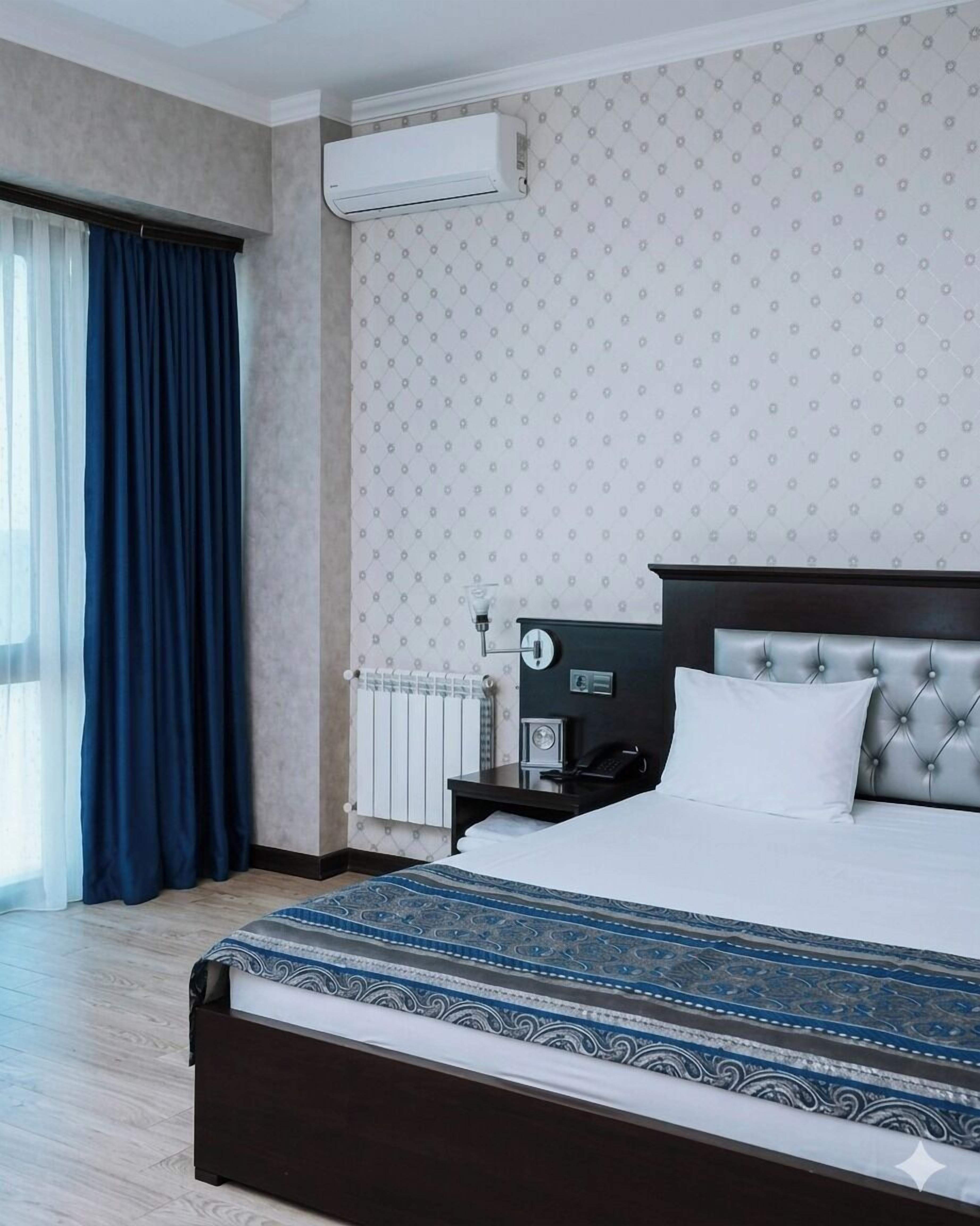 A hotel room with a large bed, a bedside table, a wall-mounted air conditioner, and a window with curtains.