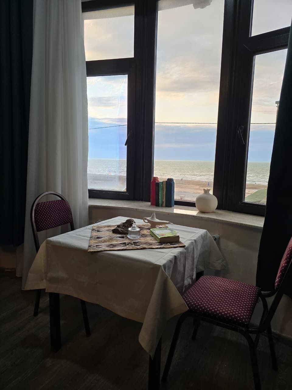 A room with a table set for two, a chair, and a view of the sea through large windows.