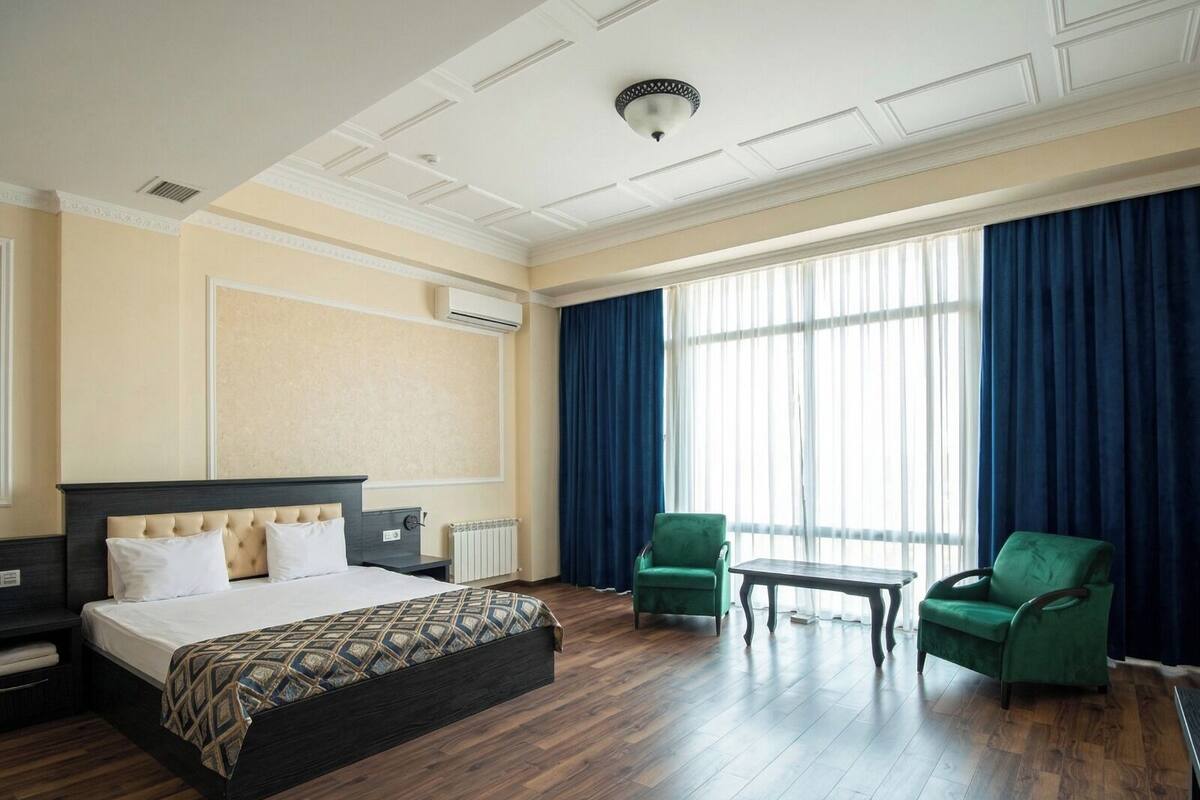 A hotel room with a large bed, two green armchairs, a small table, and a large window with curtains.