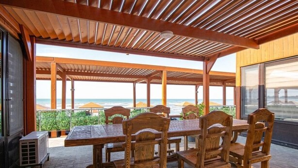 A covered outdoor dining area with wooden tables and chairs, overlooking a beach. - Gallery & Sea Hotel (Sumqayit)