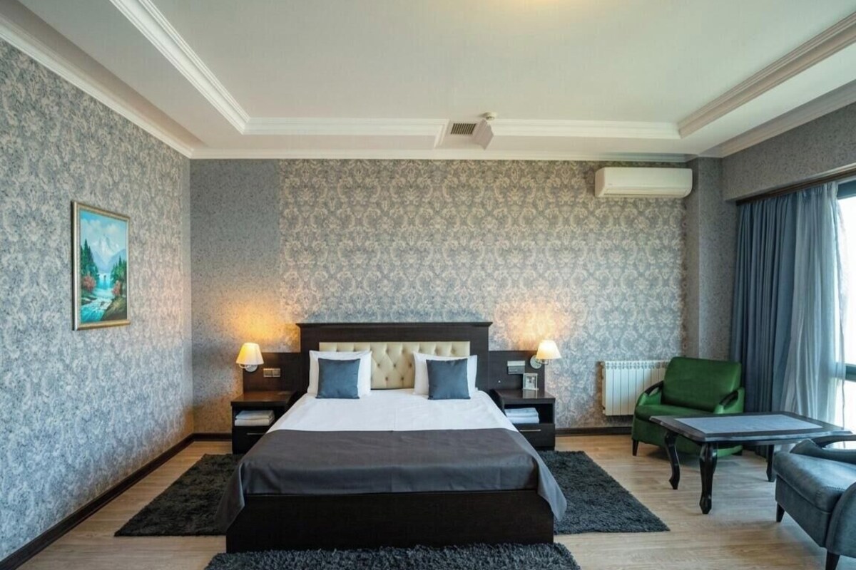 A hotel room with a large bed, two bedside tables, a green armchair, a small table, and a window with curtains.