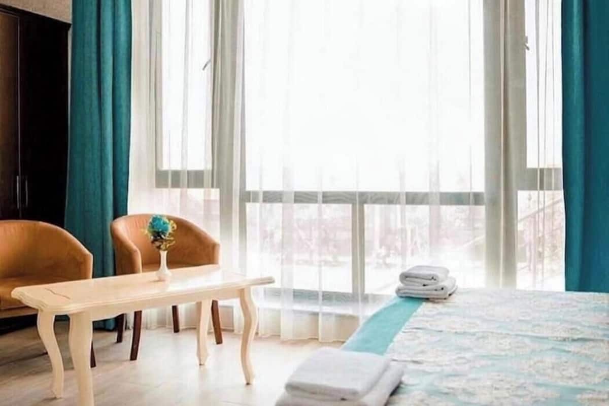A hotel room with a bed, a wooden table, two chairs, a vase, and a view through large windows.