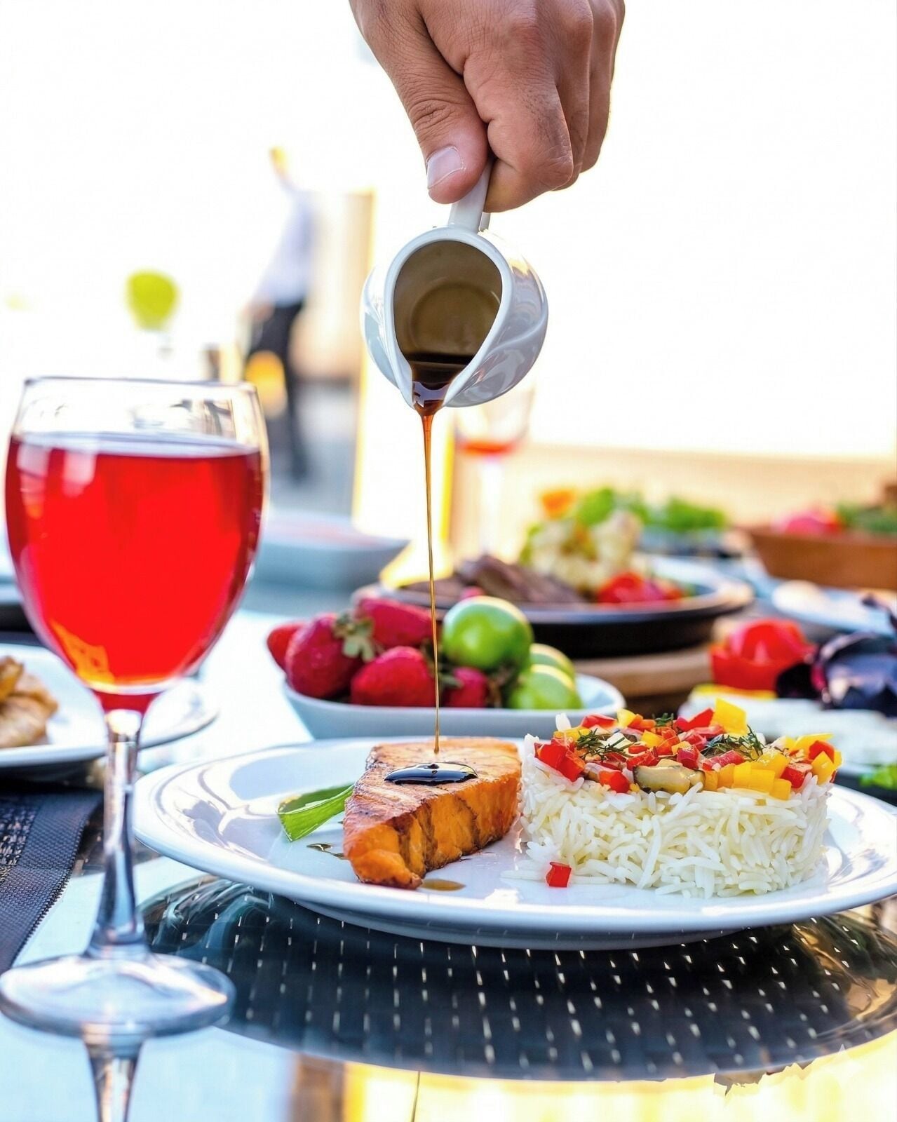 A dining table with a plate of food, a glass of red wine, and a hand pouring a sauce.