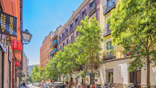 Una strada fiancheggiata da edifici a più piani, alcuni con balconi, e automobili parcheggiate lungo il marciapiede.