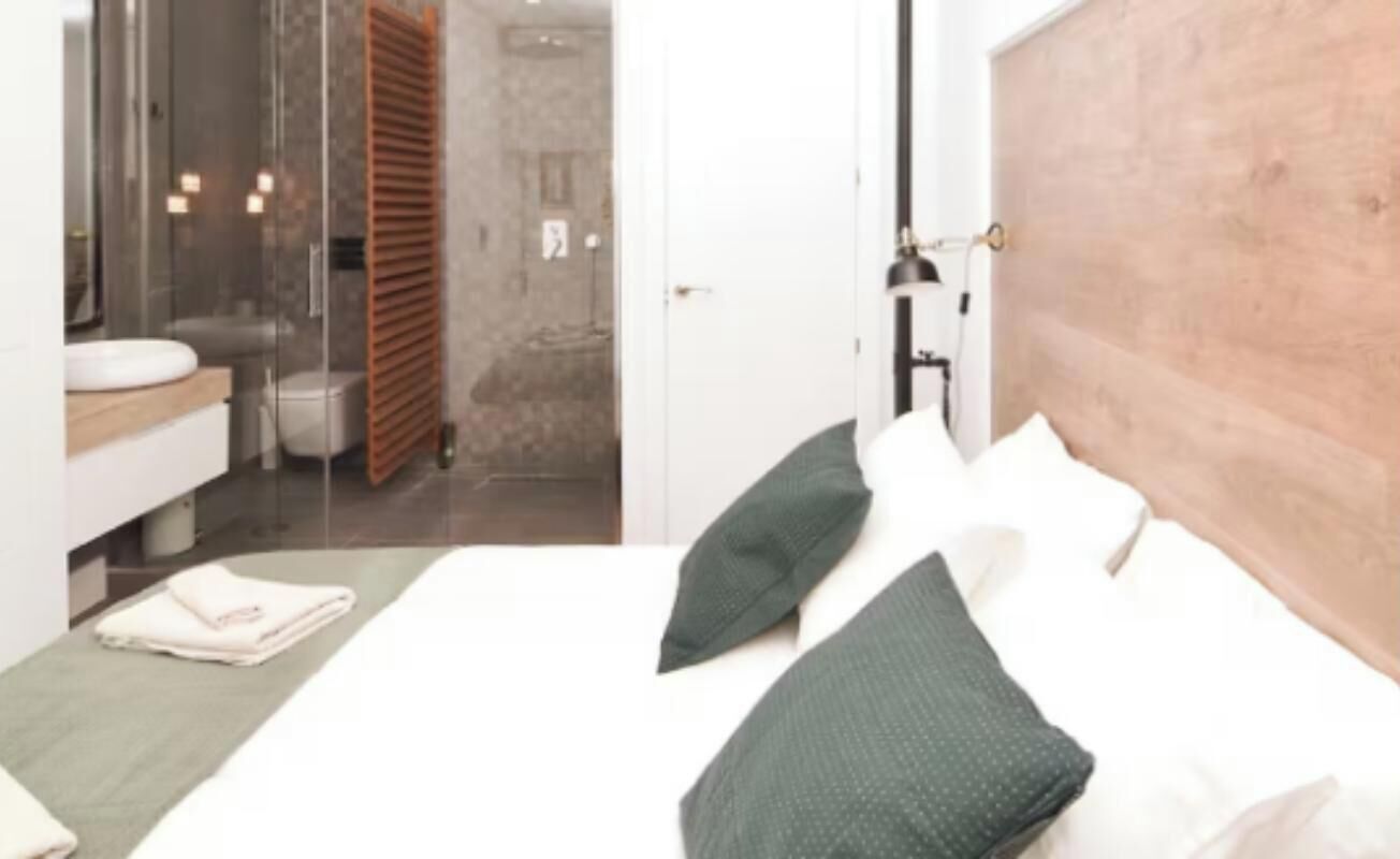 A bedroom with a wooden headboard, a bed with white and green pillows, a bedside table with a folded towel, and a view into a bathroom with a glass shower enclosure.