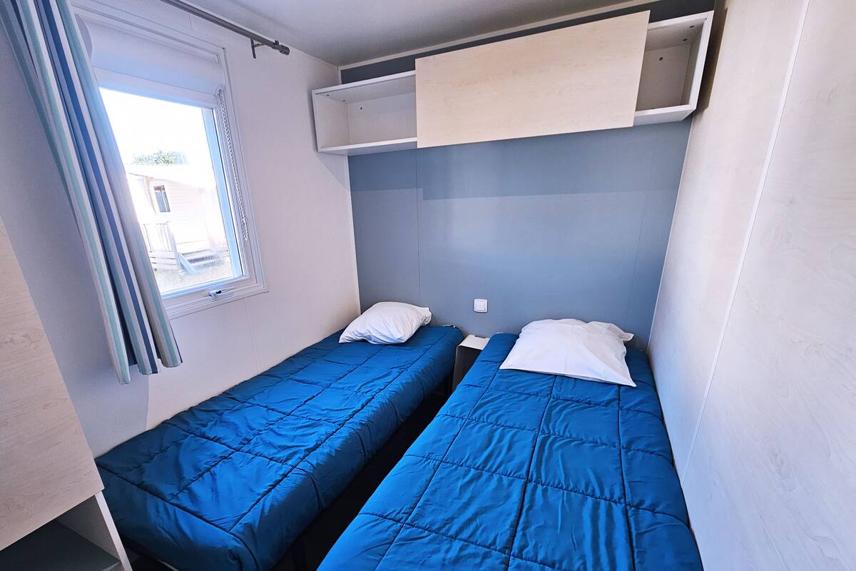 A small, compact room with two single beds, a window with curtains, and a built-in shelf above the beds.