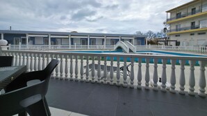 Een dakterraszwembad met een witte balustrade, een overdekt zitgedeelte en een gebouw met een blauwe gevel op de achtergrond.