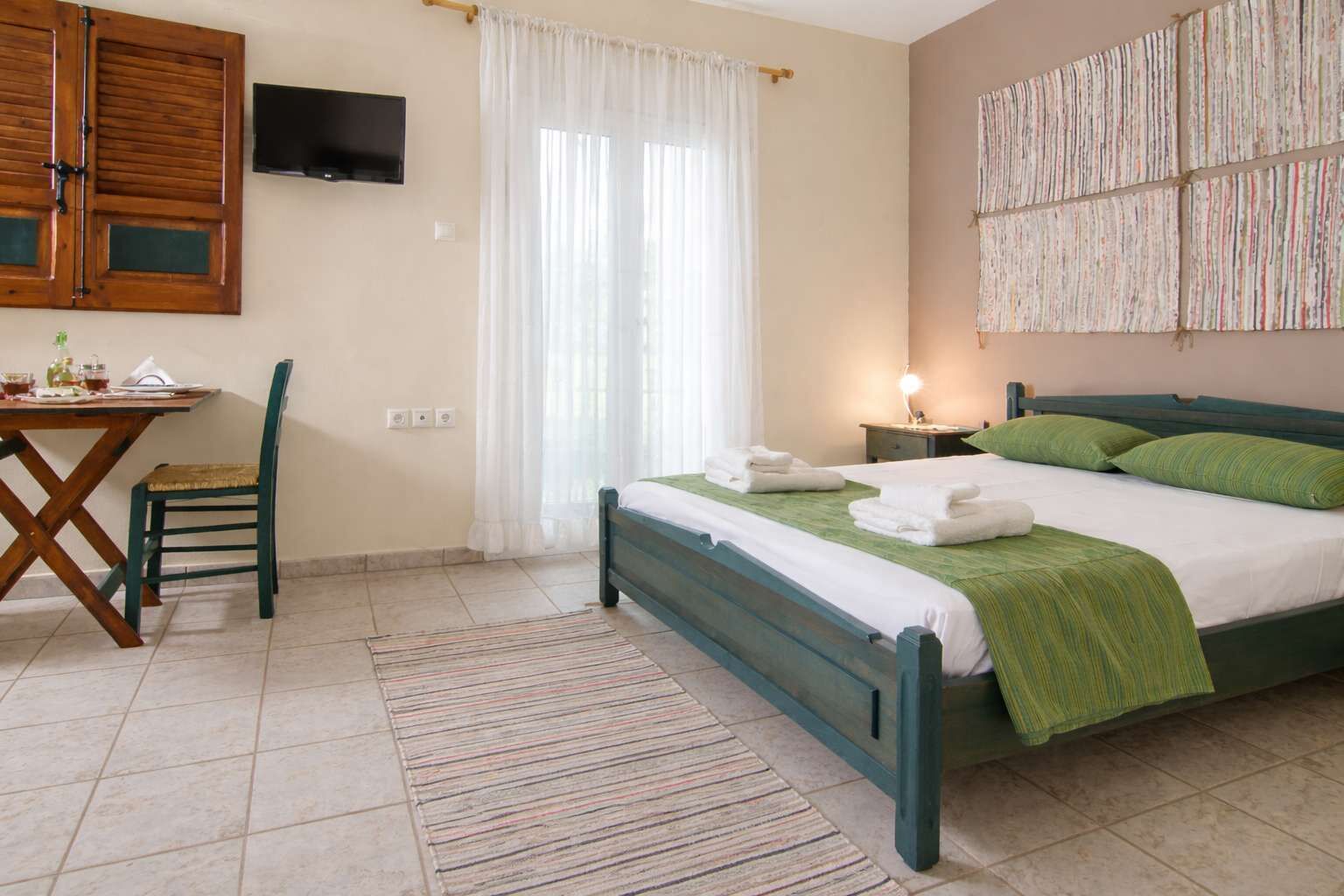 Cozy studio bedroom featuring a comfortable double bed with green linens and wooden details. The space blends rustic charm with simple decor, tiled flooring, and a calm, inviting layout ideal for a relaxing and comfortable stay