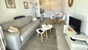 Ein modernes Wohnzimmer mit einem grauen Sofa, einem kleinen weißen Tisch, einem Fernseher und einem Essbereich mit rundem Tisch und Stühlen.