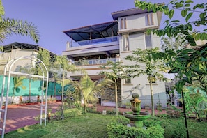 Un immeuble à plusieurs étages doté d’un balcon couvert, d’un jardin avec une fontaine et d’un ciel d’un bleu éclatant.