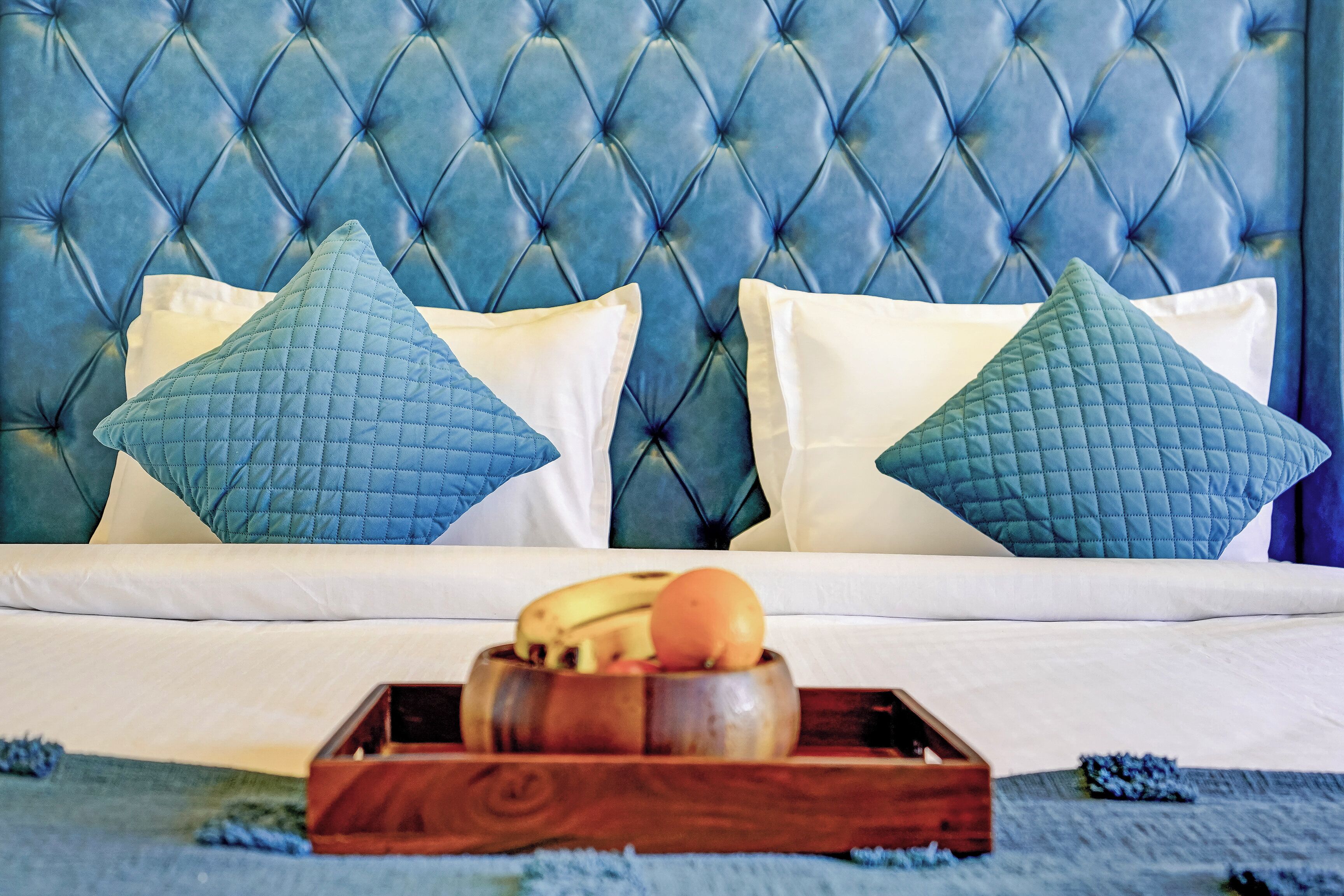 A bed with a tufted headboard, two pillows, and a wooden tray with fruits.