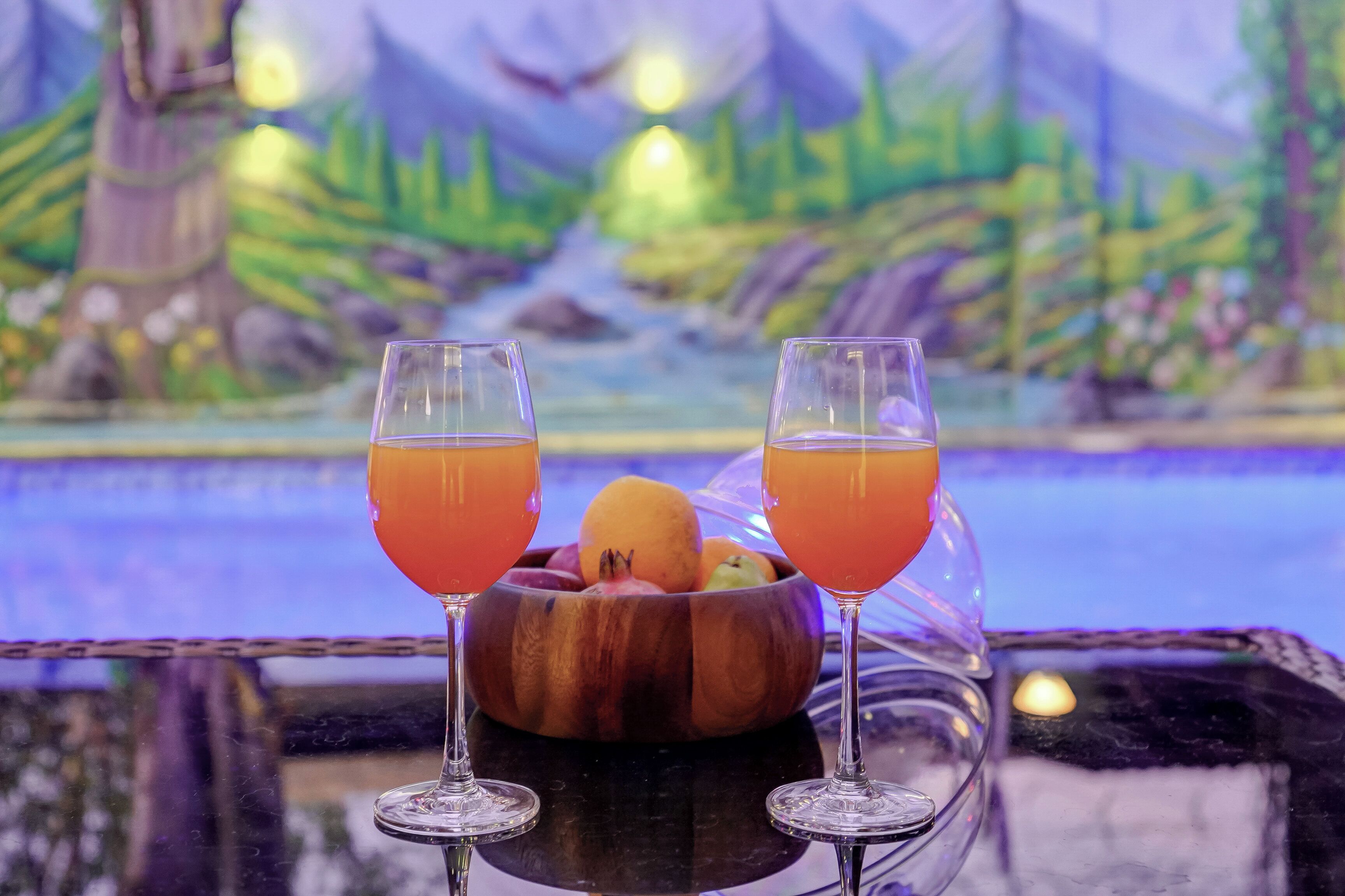 Two glasses of orange beverage on a table with a wooden bowl of fruit and a scenic mural in the background.