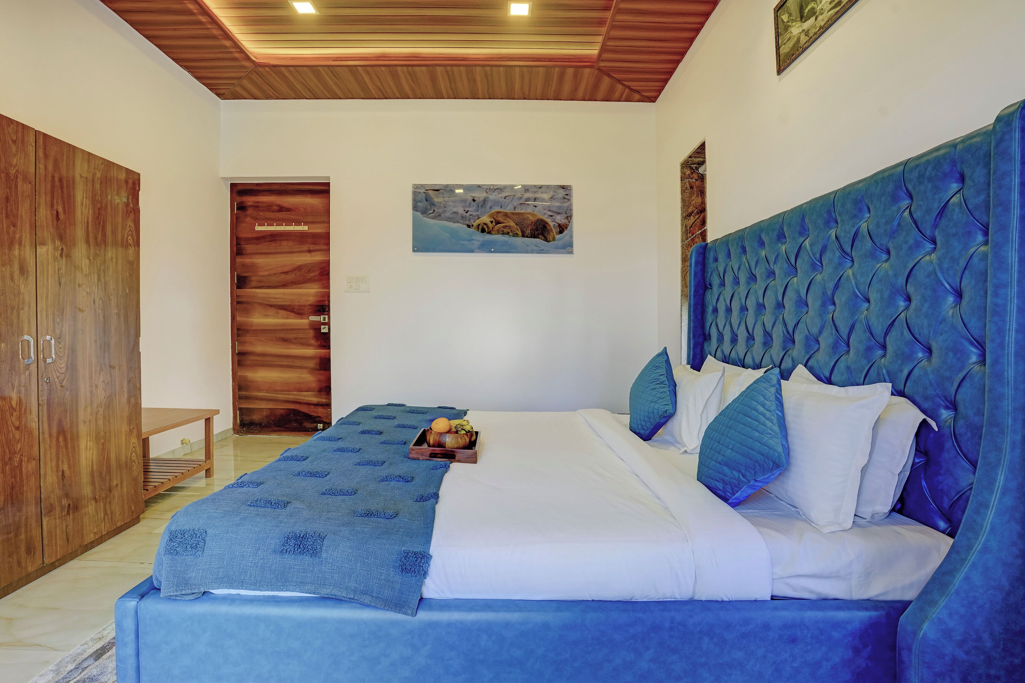 A bedroom with a bed, a wooden wardrobe, a painting on the wall, and a ceiling light.