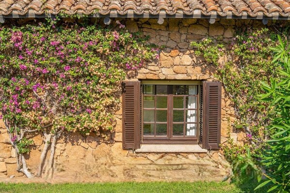 Un edificio de piedra con una ventana y contraventanas, rodeado de follaje y plantas con flores.