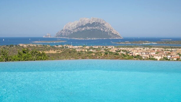 A coastal view with a mountain, clear blue water, and a town in the background. - Elite Collection Molara Infinity Pool & View (Porto San Paolo)
