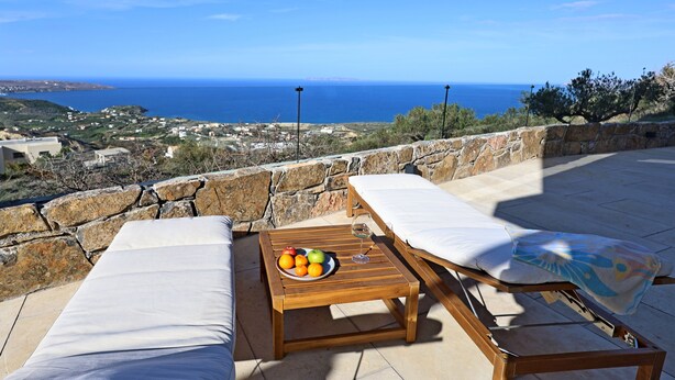 Een terras met twee houten ligstoelen, een tafel met fruit en een glas, en uitzicht op zee en de stad.