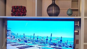 A television displaying a beach scene, a blue pot, a red decorative item, and a vase on shelves. - Luxury Apartment Very Close to the C.C. Yumbo with 2 King Size Beds 200x200cm (Maspalomas)